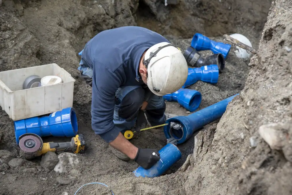 A construction worker wearing a hard hat and gloves measures a blue plastic pipe at a worksite, surrounded by pipe fittings and tools, while working in a dirt trench.